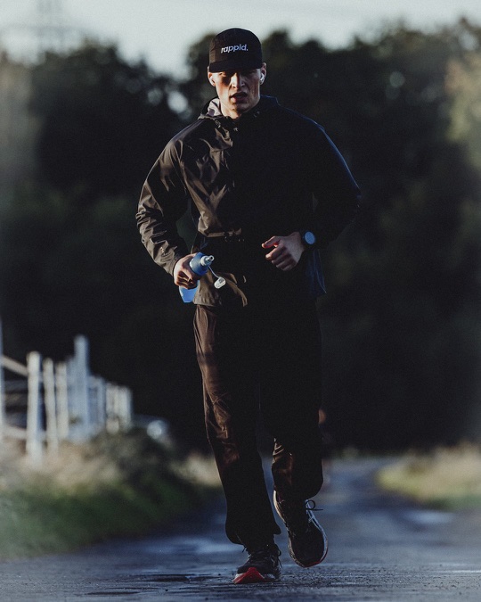 A runner in a black jacket and rappid. cap running back down a wet country road at dawn