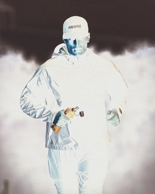 A runner in a pale jacket and rappid. cap lit by a flash against fog and darkness, carrying a soft flask